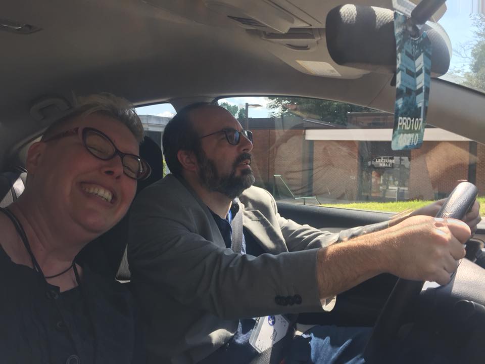 Two middle-aged white people with glasses inside a car. The man is driving, and the woman is smiling at the camera.